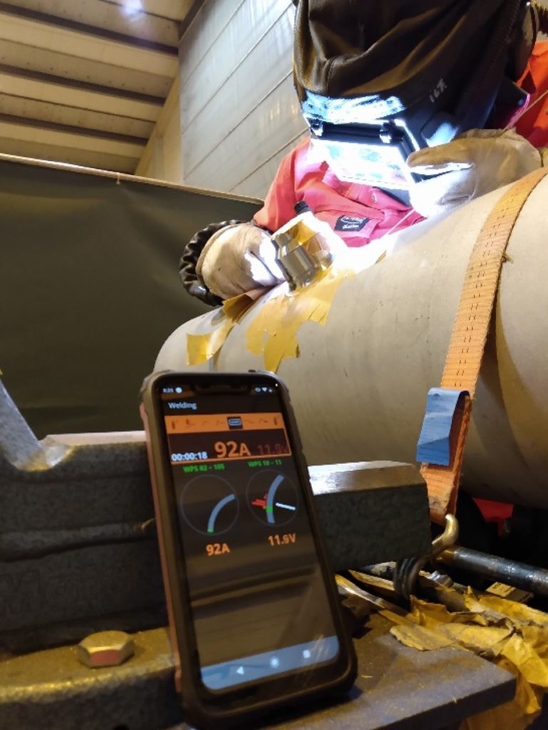 The image shows digital monitoring in a mobile phone view in front of a welder welding his work